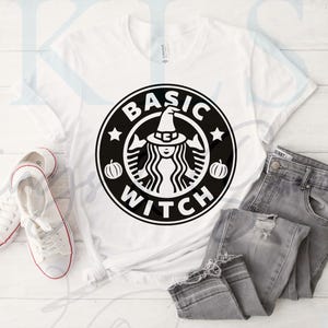 May include: White t-shirt with a black graphic reading "BASIC WITCH" in a circular design, featuring a witch with a hat, pumpkins, and stars. The shirt is paired with grey jeans and white trainers.