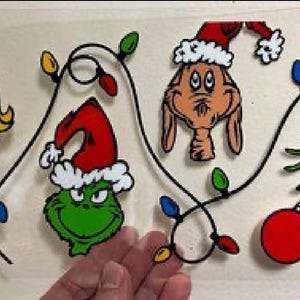 May include: A festive Christmas-themed design featuring cartoon characters and decorative elements. The design includes a smiling woman with a red bow, a green Grinch face with a Santa hat, and a brown character with a Santa hat. The design also includes a green hand holding a red ornament and string lights.
