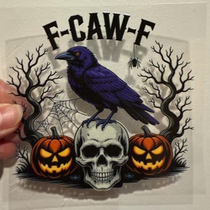 May include: A Halloween-themed graphic featuring a purple crow perched atop a skull, flanked by two jack-o'-lanterns. The design includes a spider, a spiderweb, and bare tree branches. The text "F-CAW-F" is at the top.
