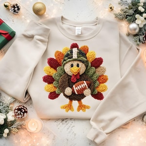 Könnte beinhalten: Cremefarbenes Sweatshirt mit einem farbenfrohen Truthahndesign. Der Truthahn trägt einen Hut und hält einen Football. Die Federn des Truthahns sind in Rot-, Gelb-, Orange- und Grüntönen gehalten. Festliche Feiertagsbekleidung.