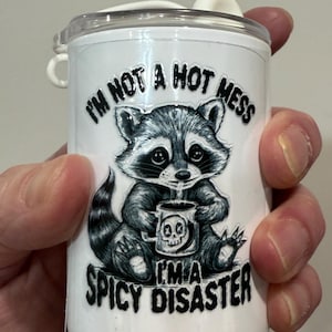 May include: White travel mug with a raccoon illustration holding a mug with a skull design. The text reads "I'm not a hot mess, I'm a spicy disaster."