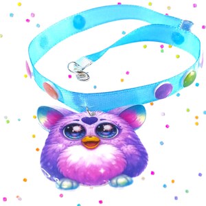 May include: A light blue pet collar with colorful polka dots and a Furby pendant. The pendant is a purple and pink Furby with large blue eyes and a yellow beak. The collar has a silver clasp.
