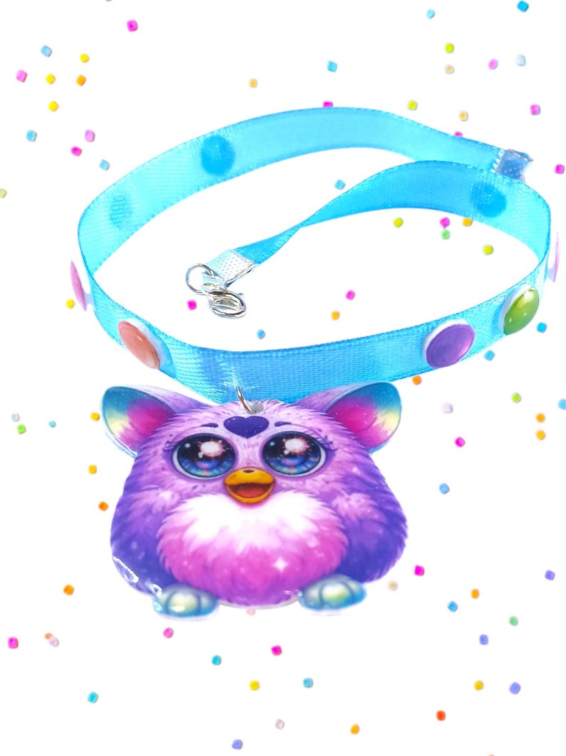 May include: A light blue pet collar with colorful polka dots and a Furby pendant. The pendant is a purple and pink Furby with large blue eyes and a yellow beak. The collar has a silver clasp.
