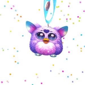 May include: A purple Furby-like charm with large, expressive eyes and a heart-shaped detail on its forehead. It has a blue ribbon with oval-shaped embellishments. Colorful confetti dots are scattered around the charm against a white background.