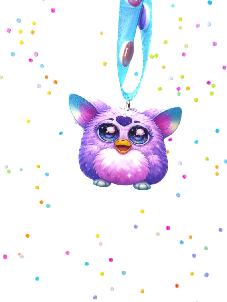 May include: A purple Furby-like charm with large, expressive eyes and a heart-shaped detail on its forehead. It has a blue ribbon with oval-shaped embellishments. Colorful confetti dots are scattered around the charm against a white background.