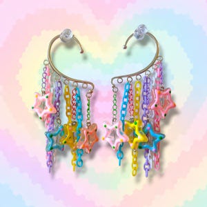 Puede incluir: Un par de puños para las orejas con un diseño de metal dorado. Cada puño presenta una barra curva con un perno transparente y cadenas colgantes en varios colores, incluyendo rosa, amarillo, azul y morado. Las cadenas están adornadas con dijes en forma de estrella.