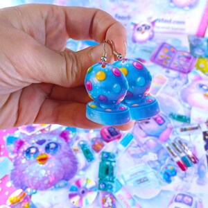 May include: A pair of blue earrings with a spherical design, featuring colorful polka dots. The earrings are held up against a background of various colorful stickers and toys, creating a playful and whimsical aesthetic.