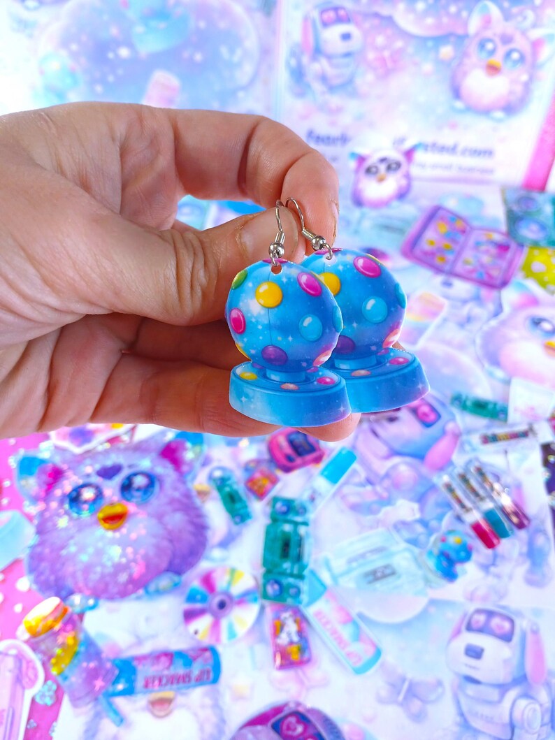 May include: A pair of blue earrings with a spherical design, featuring colorful polka dots. The earrings are held up against a background of various colorful stickers and toys, creating a playful and whimsical aesthetic.
