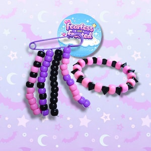 May include: A selection of handmade beaded jewellery, featuring a safety pin with pink, purple, and black beaded strands, and a bracelet with pink beads and black spikes. A round badge with the text "Fearless and Frosted" is also visible.