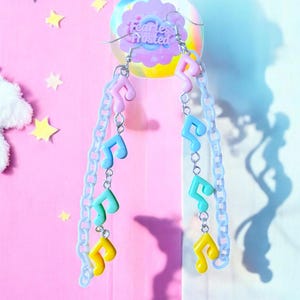 May include: A pair of pastel-coloured music note earrings with a silver chain. The earrings are hanging from a pink and blue background with the text "Fearless Frosted" on a sticker.