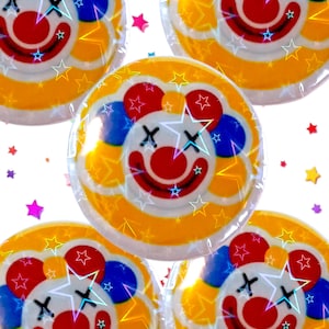 May include: Several clown-faced lollipops with a yellow, blue, and red color scheme. Each lollipop features a smiling clown face with star accents and is individually wrapped. Scattered star-shaped confetti adds a playful touch.