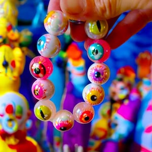 May include: A bracelet composed of spherical beads, each crafted to look like an eyeball. The beads display a variety of eye colours, such as blue, green, yellow, and red, with pink and white highlights. The bracelet is held against a bright, multi-hued backdrop.