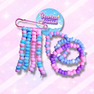 May include: A selection of colourful beaded jewellery and accessories. The image shows a pink safety pin holding strands of pink and blue beads, alongside beaded bracelets in shades of purple, pink, and blue. A blue circular badge with the text "fearless and frosted" is also present.