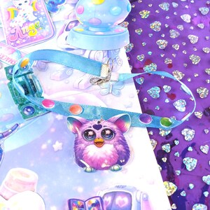 May include: A whimsical assortment of stickers and accessories. A purple Furby charm hangs from a blue ribbon. A light blue mushroom-shaped stamp and a sticker sheet with a cat are also visible. The background features a purple surface with heart-shaped glitter.
