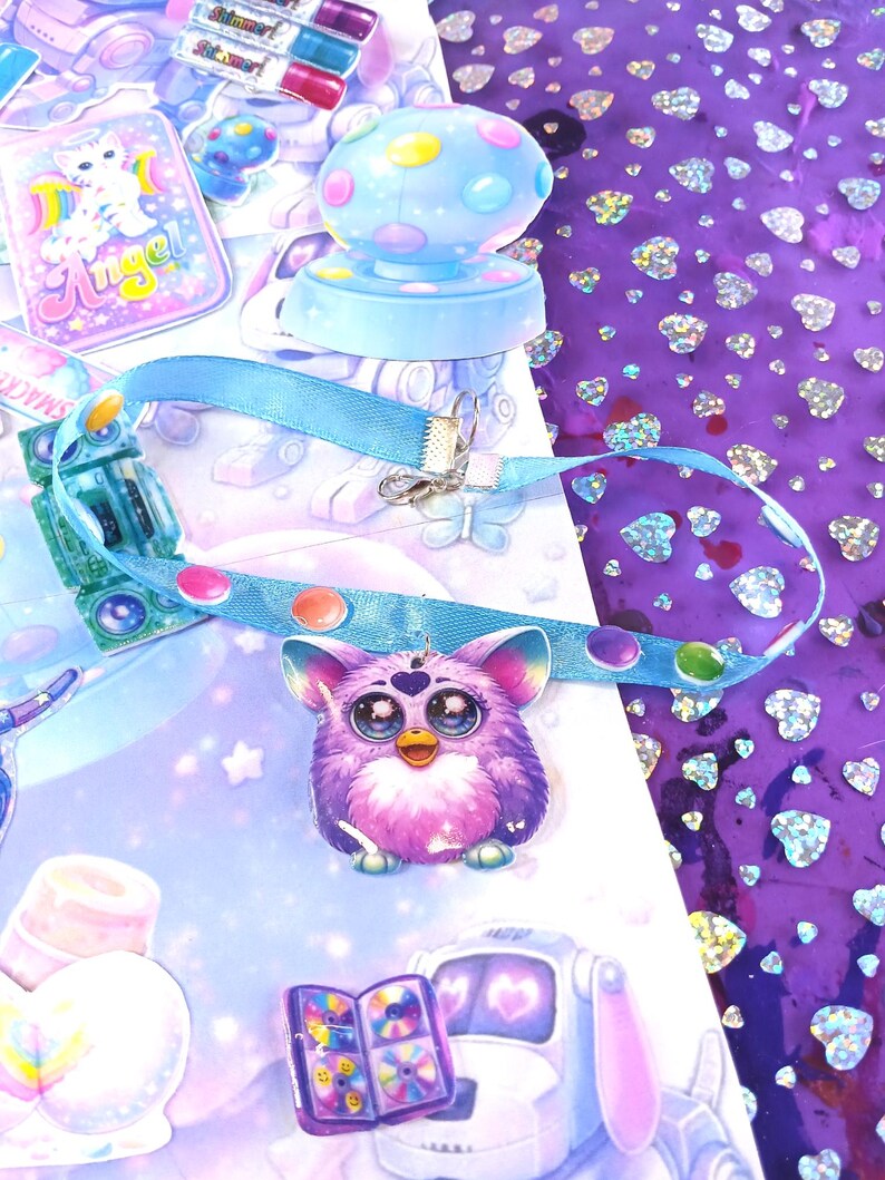 May include: A whimsical assortment of stickers and accessories. A purple Furby charm hangs from a blue ribbon. A light blue mushroom-shaped stamp and a sticker sheet with a cat are also visible. The background features a purple surface with heart-shaped glitter.