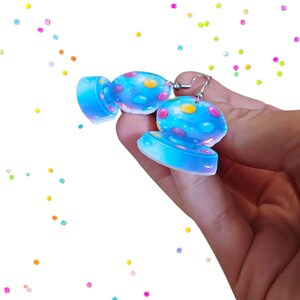 May include: A pair of blue candy-shaped earrings with colorful polka dots. The earrings are made of a translucent material and have silver-colored ear wires. The background is white with scattered colorful confetti.