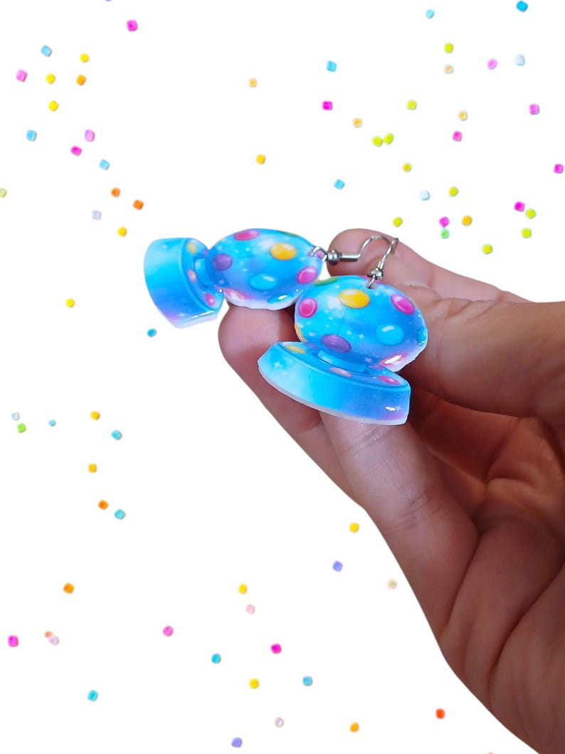 May include: A pair of blue candy-shaped earrings with colorful polka dots. The earrings are made of a translucent material and have silver-colored ear wires. The background is white with scattered colorful confetti.