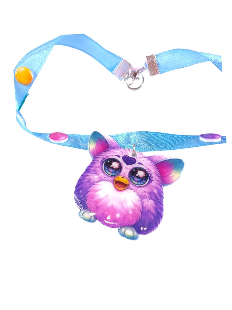 May include: A purple Furby pendant with a light blue ribbon necklace. The Furby has large, detailed eyes and a pink and purple body. The ribbon has decorative buttons and a metal clasp.