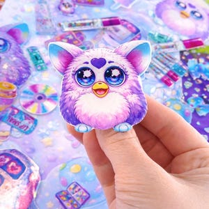 Könnte beinhalten: Ein farbenfroher Aufkleber mit einem Furby-ähnlichen Charakter mit großen blauen Augen, einem lila Herzen und einem gelben Schnabel. Der Aufkleber befindet sich vor einem Hintergrund aus anderen Aufklebern und Designs in Lila-, Rosa- und Blautönen.
