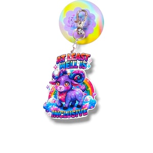 May include: A colorful keychain featuring a purple ram with large horns, a rainbow, and the text "AT LEAST HELL IS INCLUSIVE". The keychain is attached to a round, rainbow-colored stress ball with a silver clasp.