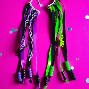 May include: Two decorative ear cuffs with white hooks, black, purple, and green ribbons, chains, and beads. The ear cuffs are set against a bright pink background with silver confetti.