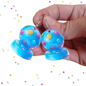 May include: A pair of blue, dome-shaped earrings with colorful polka dots and silver hooks. The earrings have a base and a rounded top, with a starry sky design. The earrings are held in a hand against a white background with colorful confetti.