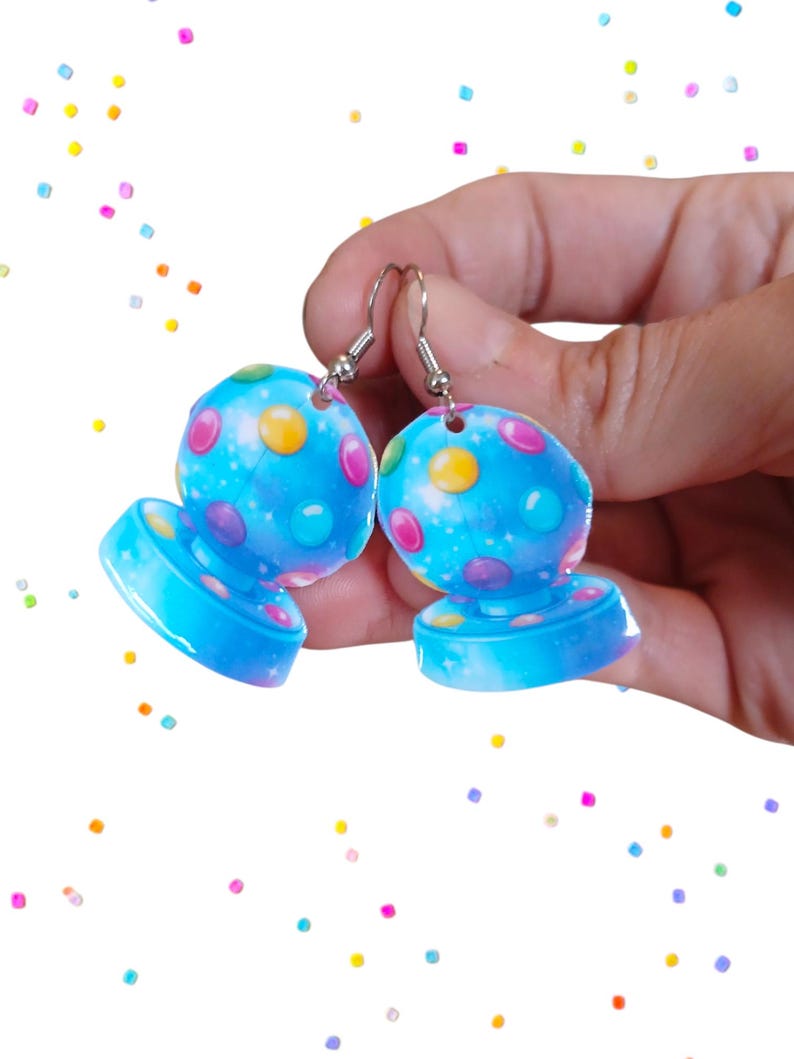 May include: A pair of blue, dome-shaped earrings with colorful polka dots and silver hooks. The earrings have a base and a rounded top, with a starry sky design. The earrings are held in a hand against a white background with colorful confetti.
