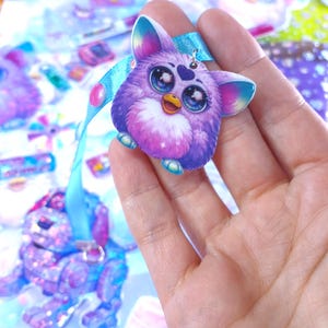 May include: A purple Furby-inspired charm with large blue eyes, a yellow beak, and a heart-shaped detail. It is attached to a light blue ribbon. The charm is held in a hand, with other similar charms visible in the background.