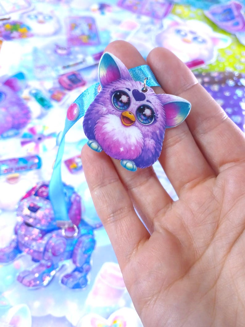 May include: A purple Furby-inspired charm with large blue eyes, a yellow beak, and a heart-shaped detail. It is attached to a light blue ribbon. The charm is held in a hand, with other similar charms visible in the background.