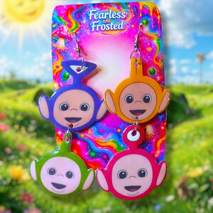 May include: Colorful earrings featuring four cartoon characters in purple, orange, green, and pink. The earrings are attached to a card with the text "Fearless and Frosted" on a psychedelic background. The background is a sunny day with a blue sky and green grass.