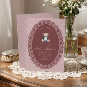 May include: A light pink baby shower invitation card featuring a dark red oval frame with a lace-like border. The center displays a teddy bear illustration and text reading A LITTLE FIRECRACKER IS ON THE WAY PLEASE JOIN US FOR A BABY SHOWER.