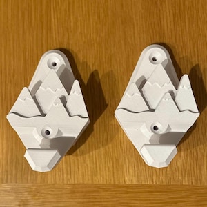 Puede incluir: Dos ganchos de pared blancos impresos en 3D con forma de montañas. Cada gancho tiene un agujero para colgar objetos y un pequeño detalle en relieve de una cordillera.