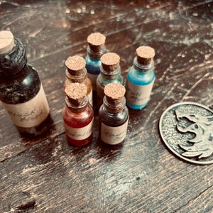 Pode incluir: Uma coleção de seis pequenas garrafas de vidro com rolhas de cortiça, contendo vários líquidos coloridos e rotuladas com texto manuscrito, ao lado de um pingente circular de metal prateado com um emblema de cabeça de lobo.
