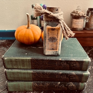 Pode incluir: Uma garrafa de vidro quadrada com rolha de cortiça e um laço de corda de juta, com um rótulo retangular com texto, sobre uma pilha de três livros antigos verde-escuros ao lado de uma pequena abóbora laranja e outros frascos.