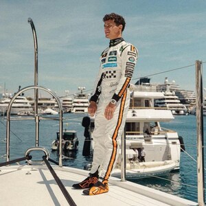 May include: A person wearing a white racing suit with orange stripes and black accents, standing on a yacht. The suit features sponsor logos and a checkered pattern. The person is wearing a watch and black and orange shoes. The background includes boats and the sea.