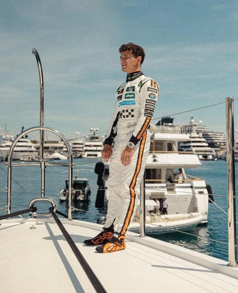 May include: A person wearing a white racing suit with orange stripes and black accents, standing on a yacht. The suit features sponsor logos and a checkered pattern. The person is wearing a watch and black and orange shoes. The background includes boats and the sea.