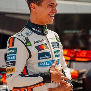 May include: A smiling person wearing a white racing suit with black and orange accents. The suit has sponsor logos including "Android", "DP WORLD", and "DELL Technologies". The number "4" is visible on the suit.