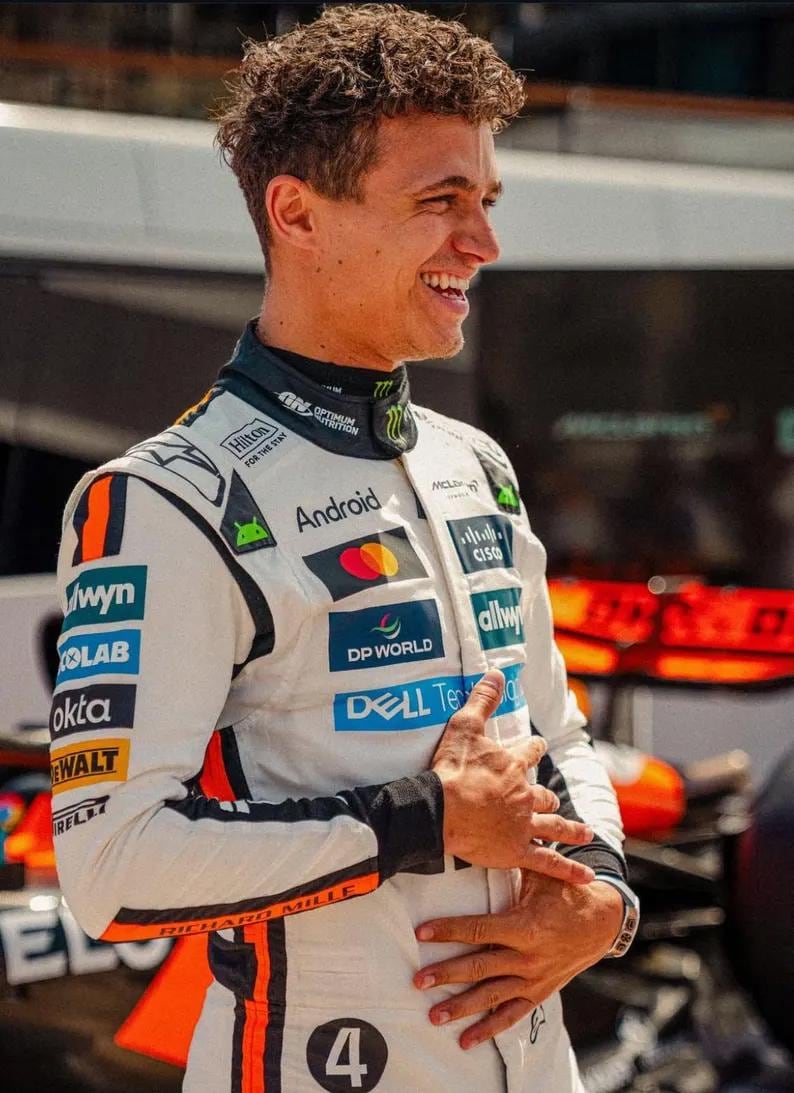 May include: A smiling person wearing a white racing suit with black and orange accents. The suit has sponsor logos including "Android", "DP WORLD", and "DELL Technologies". The number "4" is visible on the suit.