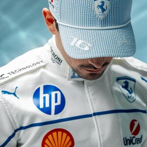 May include: A light blue and white striped baseball cap with the number "16" on the side and a Ferrari logo. The person is wearing a white racing suit with blue accents and sponsor logos, including HP, Shell, and UniCredit.