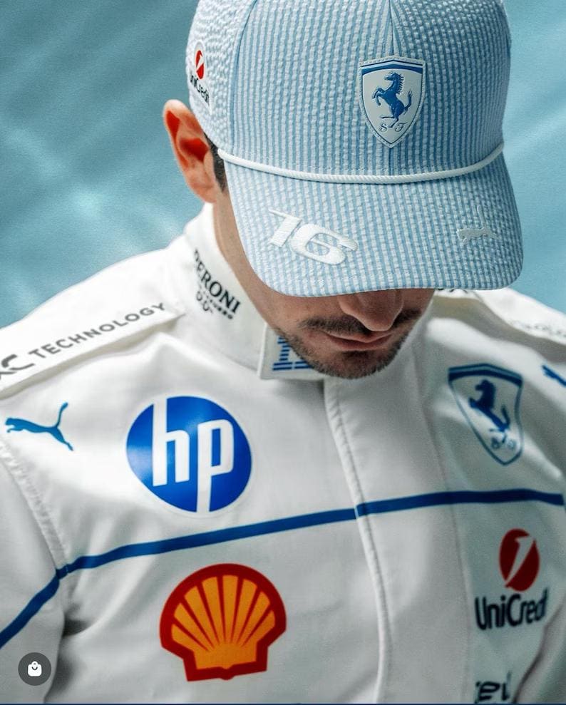 May include: A light blue and white striped baseball cap with the number "16" on the side and a Ferrari logo. The person is wearing a white racing suit with blue accents and sponsor logos, including HP, Shell, and UniCredit.