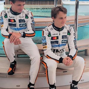 May include: Two people wearing white racing suits with black and orange accents, sitting on a wooden surface. The suits feature sponsor logos, including "Android," "Cisco," and "McLaren." They are wearing black and orange racing shoes. The background shows a body of water.