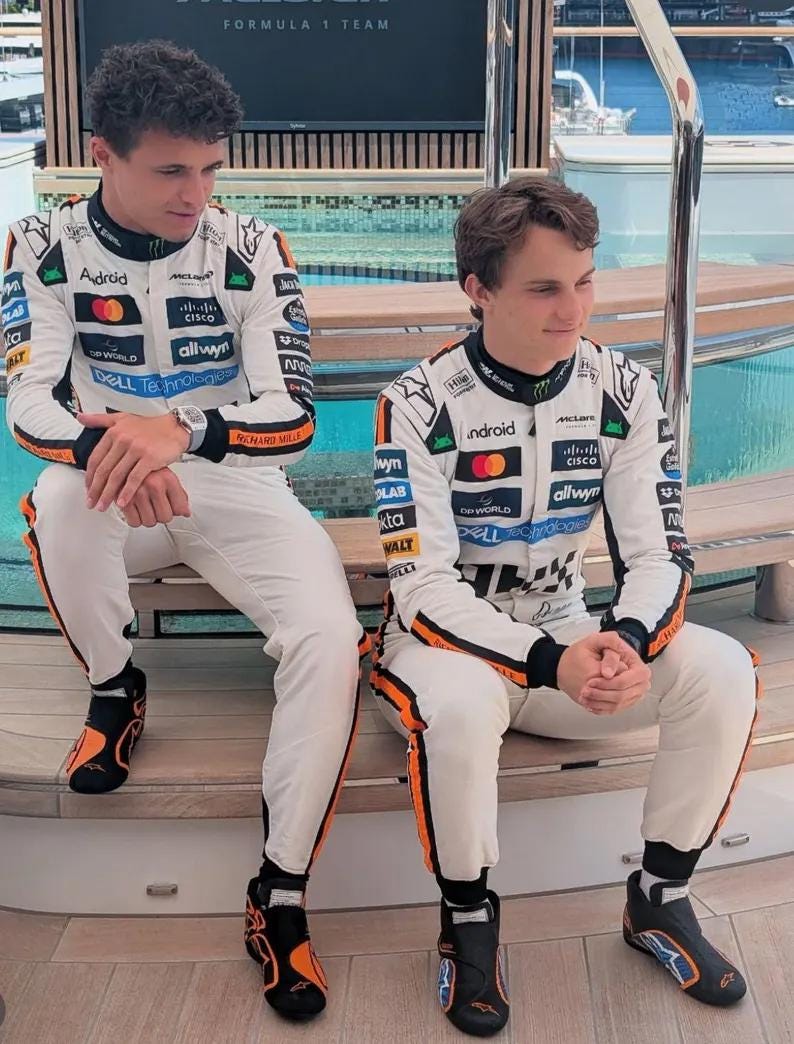 May include: Two people wearing white racing suits with black and orange accents, sitting on a wooden surface. The suits feature sponsor logos, including "Android," "Cisco," and "McLaren." They are wearing black and orange racing shoes. The background shows a body of water.