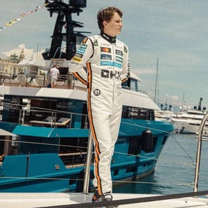 May include: A person wearing a white racing suit with orange stripes and sponsor logos stands on a yacht. The suit has the number 81 on the chest. The background includes a blue yacht and the ocean.
