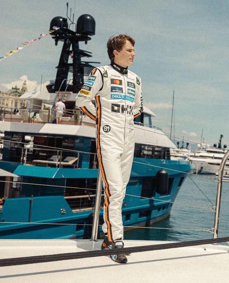 May include: A person wearing a white racing suit with orange stripes and sponsor logos stands on a yacht. The suit has the number 81 on the chest. The background includes a blue yacht and the ocean.
