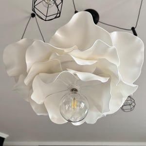 May include: A large, white, flower-shaped ceiling light fixture. The fixture has multiple layers of petal-like shapes and a clear lightbulb in the center. Additional black, geometric pendant lights are visible in the background.