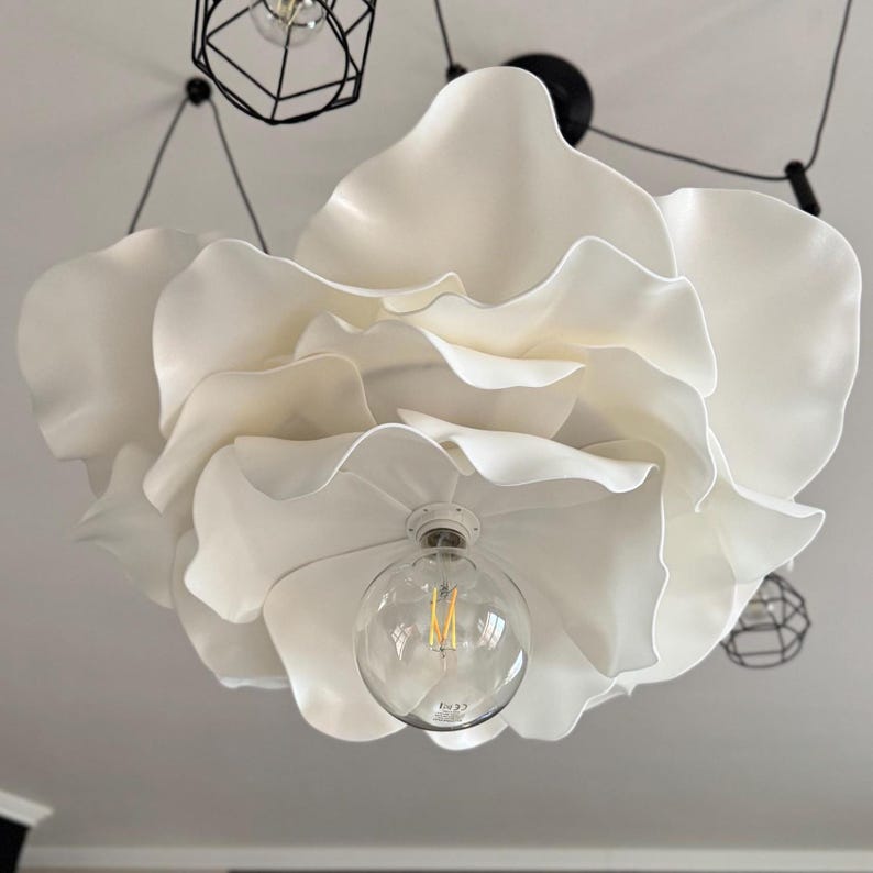 May include: A large, white, flower-shaped ceiling light fixture. The fixture has multiple layers of petal-like shapes and a clear lightbulb in the center. Additional black, geometric pendant lights are visible in the background.