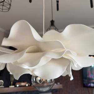 May include: A large, white, flower-shaped pendant light fixture. The light has multiple layers of petal-like shades, creating a soft, diffused glow. A spherical glass bulb hangs below the flower. The fixture is suspended from the ceiling.