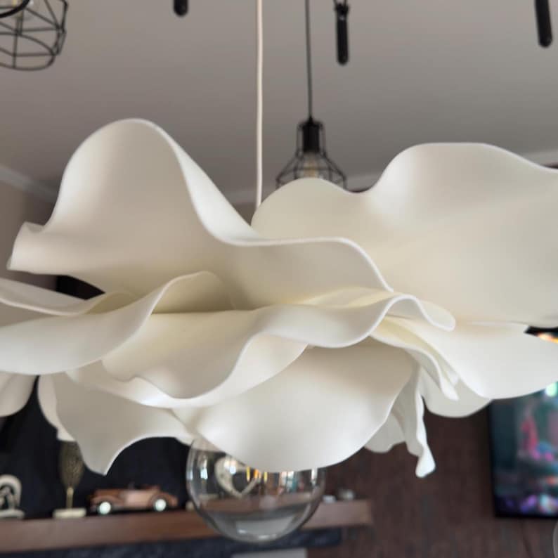 May include: A large, white, flower-shaped pendant light fixture. The light has multiple layers of petal-like shades, creating a soft, diffused glow. A spherical glass bulb hangs below the flower. The fixture is suspended from the ceiling.
