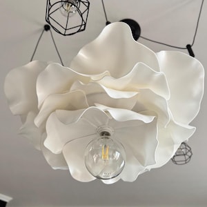 May include: A large, white, flower-shaped ceiling light fixture. The light has multiple layers of petal-like shades and a clear glass bulb. Additional black geometric pendant lights are visible in the background.