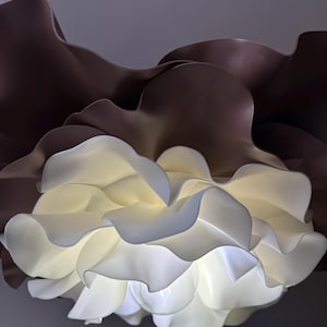 Può includere: Una grande scultura floreale astratta con petali a strati. I petali esterni sono marrone scuro, mentre i petali interni sono di un bianco brillante, creando un contrasto sorprendente. La scultura è illuminata dall'interno, conferendole una morbida lucentezza.